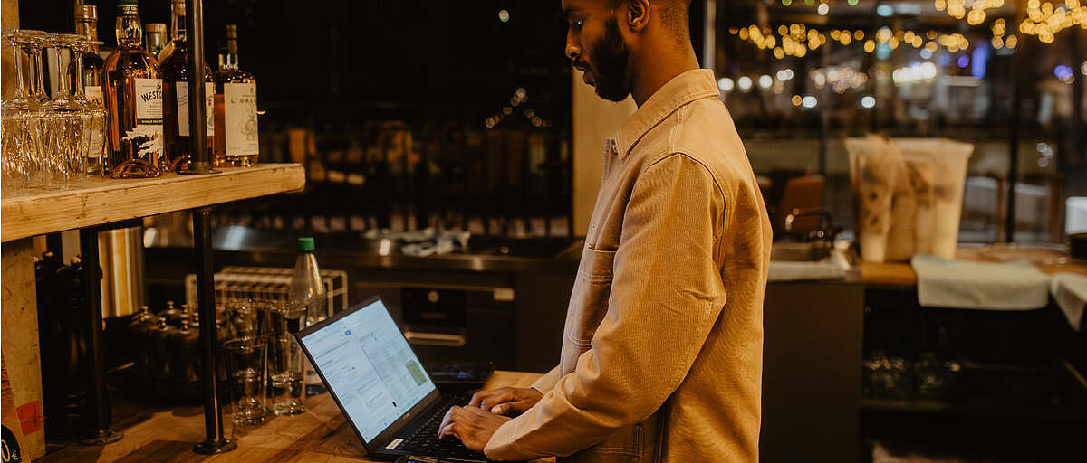 Mitarbeiter im Hopfenfänger Brauhaus arbeitet an einem Laptop an der Bar, im Hintergrund Gläser und Flaschen