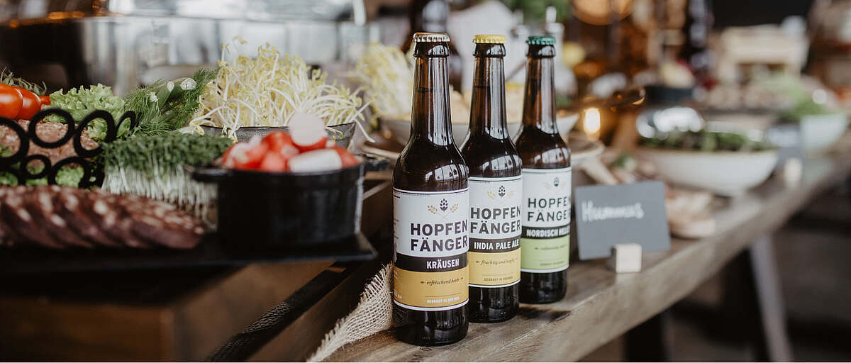 Drei Hopfenfänger Bierflaschen – Kräusen, India Pale Ale und Nordisch Herb – auf einem Catering-Buffet mit frischen Zutaten