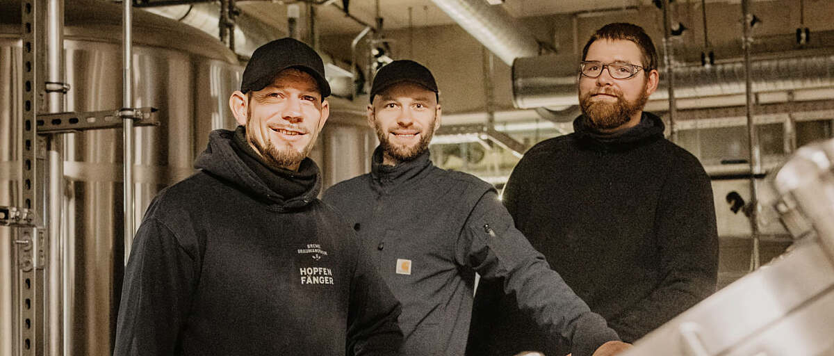 Drei Brauer des Hopfenfänger Teams in der Brauerei, vor Edelstahlkesseln und Brauanlage
