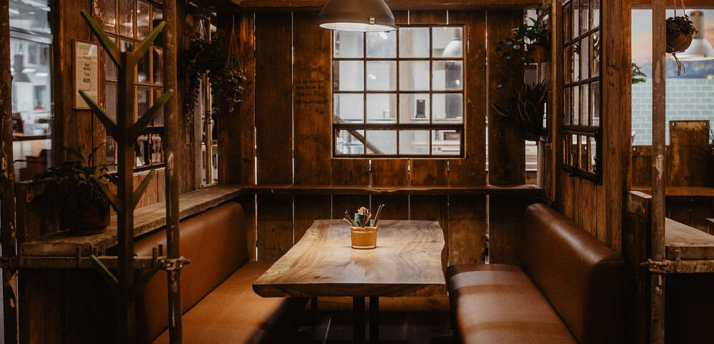 Gemütliche Sitzecke im Hopfenfänger Restaurant mit Holztisch, braunen Ledersofas, Pflanzen und rustikaler Holzwand mit Fenstern