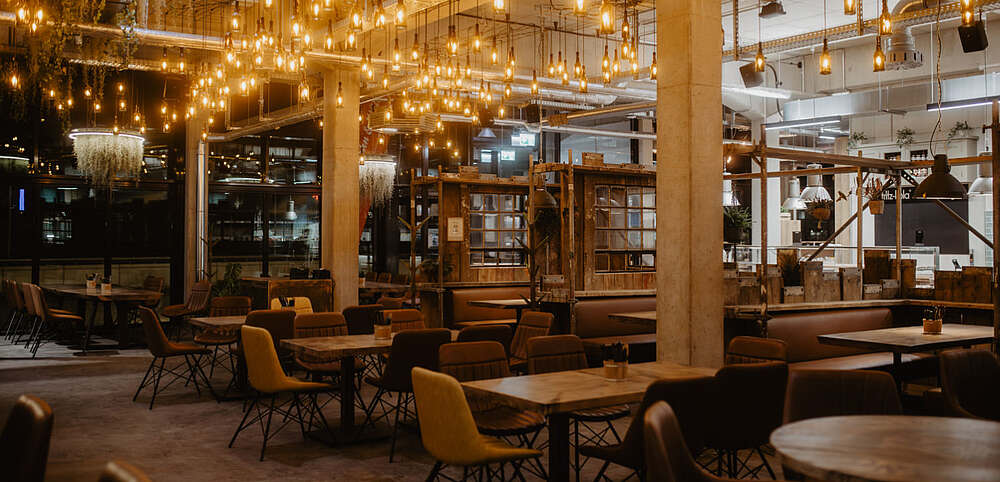 Innenraum des stimmungsvoll beleuchteten Restaurants/Brauereigaststätte mit rustikalen Holztischen, gemütlichen Stühlen und zahlreichen Hängelampen
