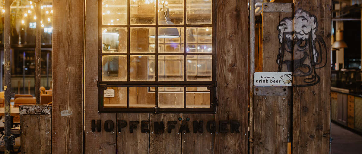 Holzwand mit Fenster im Hopfenfänger Brauhaus, dekoriert mit dem Schriftzug Hopfenfänger und Bierkrug-Grafik