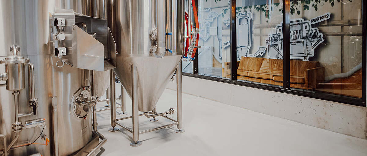 Edelstahl-Gärtanks in der Hopfenfänger Brauerei, dahinter Glaswand mit Illustration des Brauprozesses und einer Ledercouch im Besucherbereich
